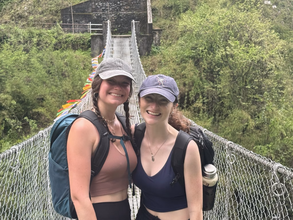 Two friends smiling on a suspension bridge covered with prayer flags during their Pokhara trek in Nepal.